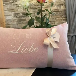Zirbenkissen Geschenk zum Geburtstag. Zirbenkissen mit Baumwollüberzug in Farbe rosé. Auf dem Kissenüberzug ist eine gestickte Personalisierung in Farbe creme zu sehen. Die Personalisierung lautet DU BIST Liebe. Eine cremefarbige Satinbandschleife ist auch auf der Abbildung zu sehen.